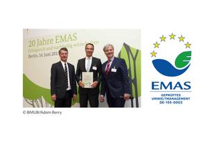 Drei Männer im Anzug bei einer EMAS-Jubiläumsveranstaltung, einer hält eine Urkunde, daneben das EMAS-Umweltmanagement-Logo.