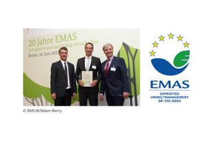 Drei Männer im Anzug bei einer EMAS-Jubiläumsveranstaltung, einer hält eine Urkunde, daneben das EMAS-Umweltmanagement-Logo.