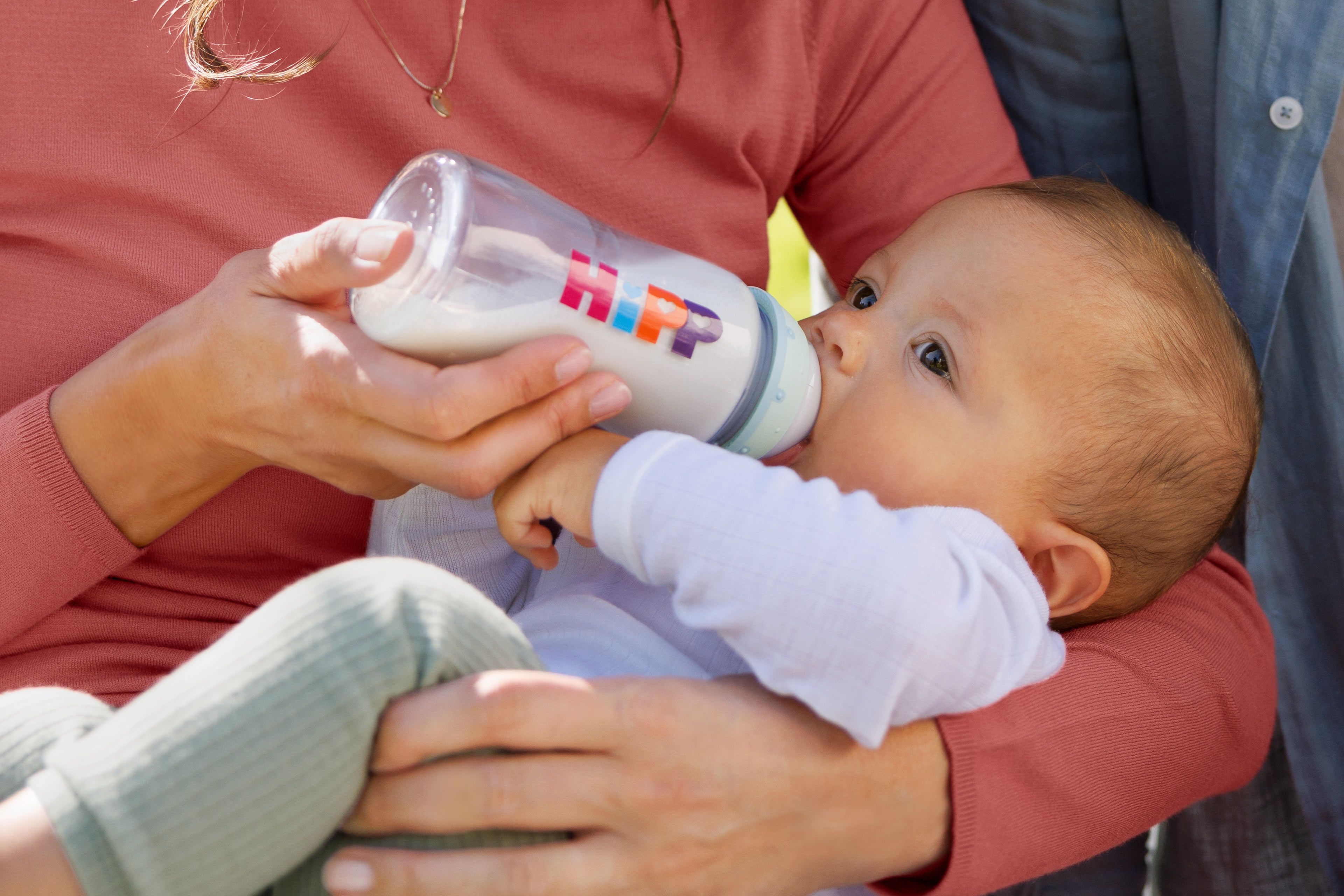 Baby wird von einer Person im roten Pullover mit einer Flasche HiPP-Milch gefüttert