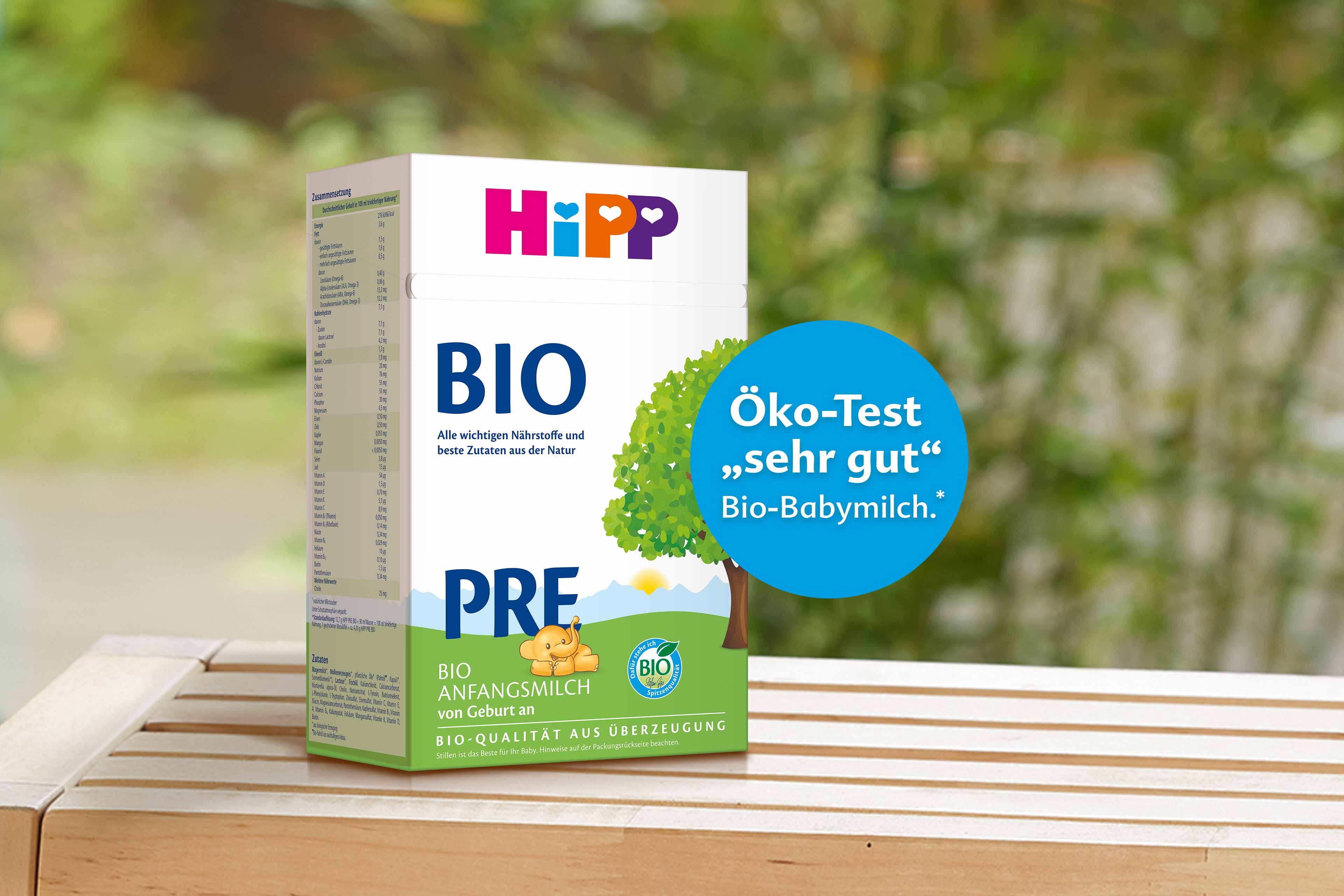 Packung HiPP BIO Pre Anfangsmilch auf Holztisch, daneben blauer Kreis mit Aufdruck „Öko-Test ‚sehr gut‘ Bio-Babymilch“ vor Naturhintergrund.