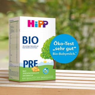 Packung HiPP BIO Pre Anfangsmilch auf Holztisch, daneben blauer Kreis mit Aufdruck „Öko-Test ‚sehr gut‘ Bio-Babymilch“ vor Naturhintergrund.