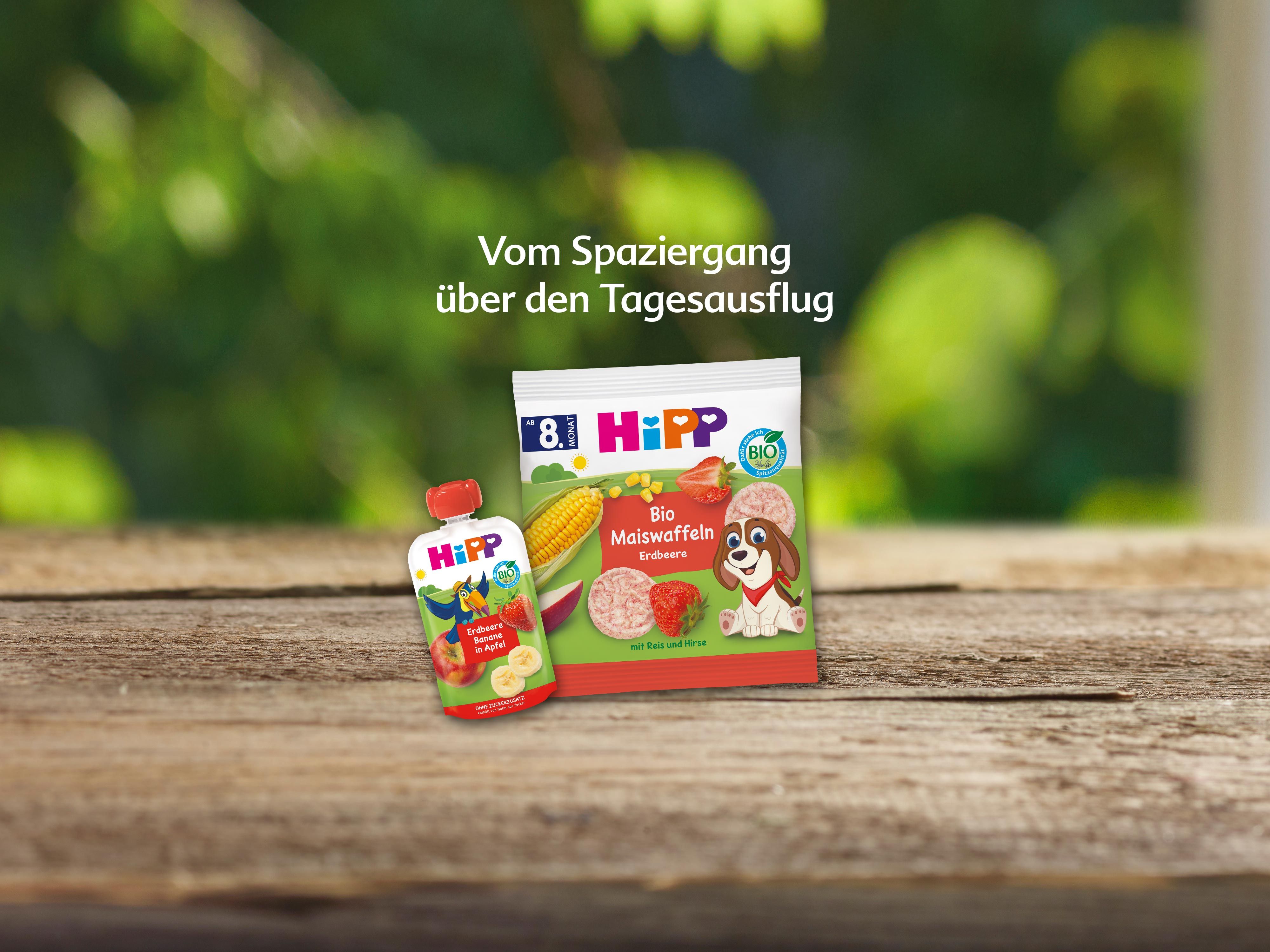 Packung Bio-Maiswaffeln und Quetschbeutel mit Erdbeergeschmack von HiPP auf Holztisch vor grünem Hintergrund