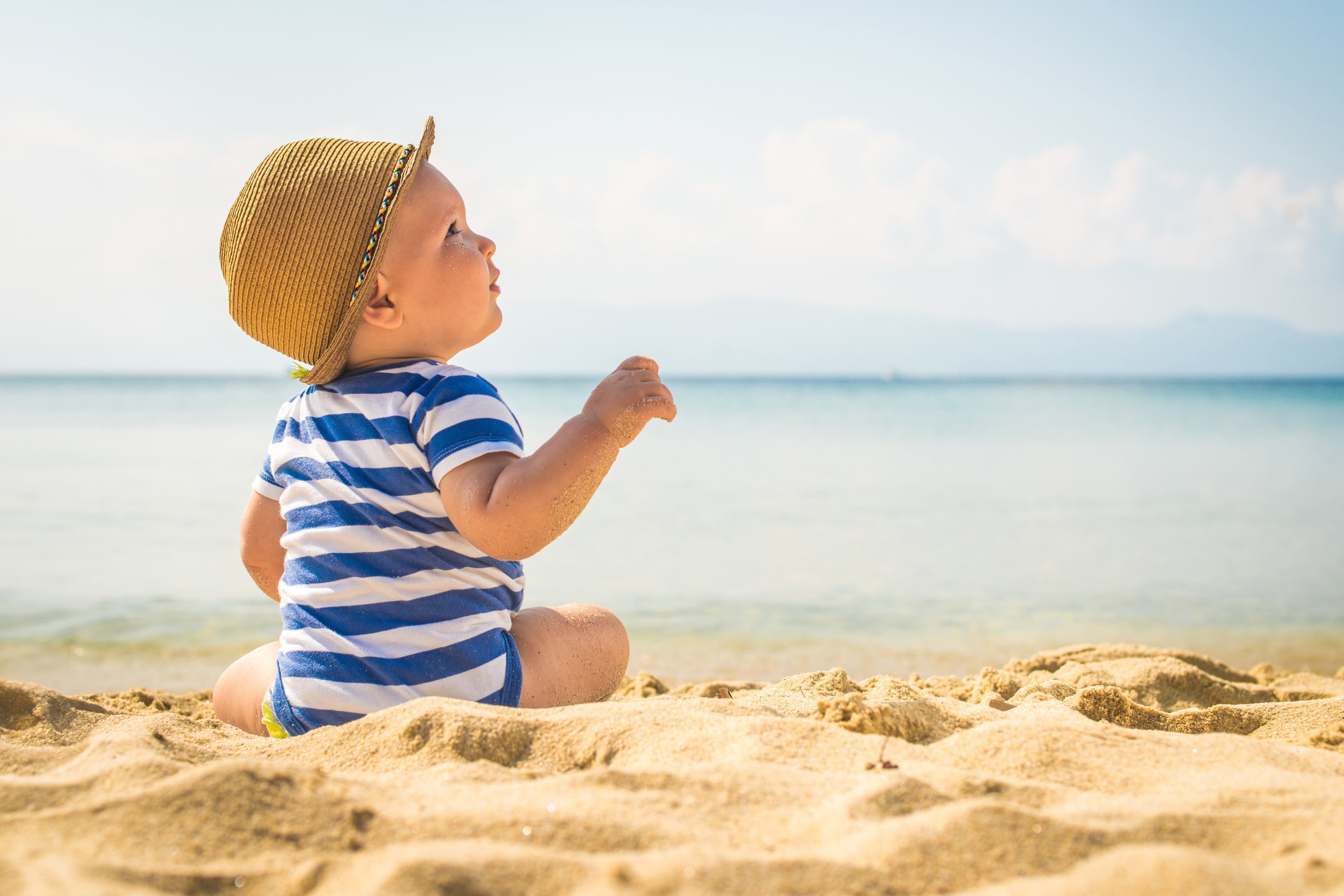 Ein kleines Kind in einem gestreiften Outfit und mit einem Hut sitzt an einem Sandstrand und blickt in die Ferne mit einer ruhigen Kulisse des Meeres und der fernen Berge.