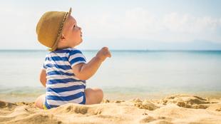 Ein kleines Kind in einem gestreiften Outfit und mit einem Hut sitzt an einem Sandstrand und blickt in die Ferne mit einer ruhigen Kulisse des Meeres und der fernen Berge.