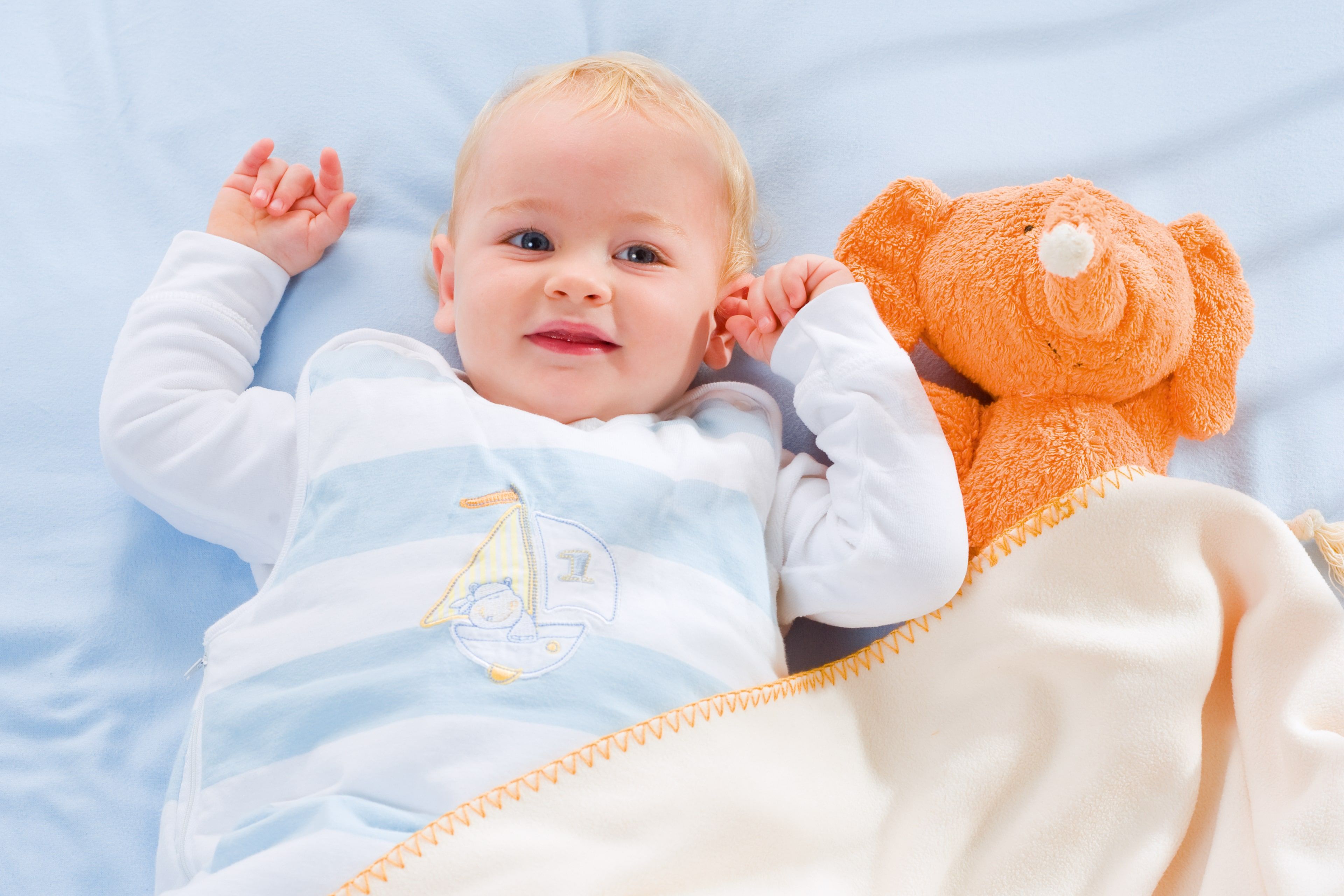 Ein Baby, das auf einer blauen Fläche liegt und ein weißes Outfit mit einem Segelbootmuster trägt, neben einem orangefarbenen Plüschtier, das einem Elefanten ähnelt.