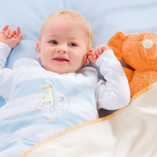 Ein Baby, das auf einer blauen Fläche liegt und ein weißes Outfit mit einem Segelbootmuster trägt, neben einem orangefarbenen Plüschtier, das einem Elefanten ähnelt.