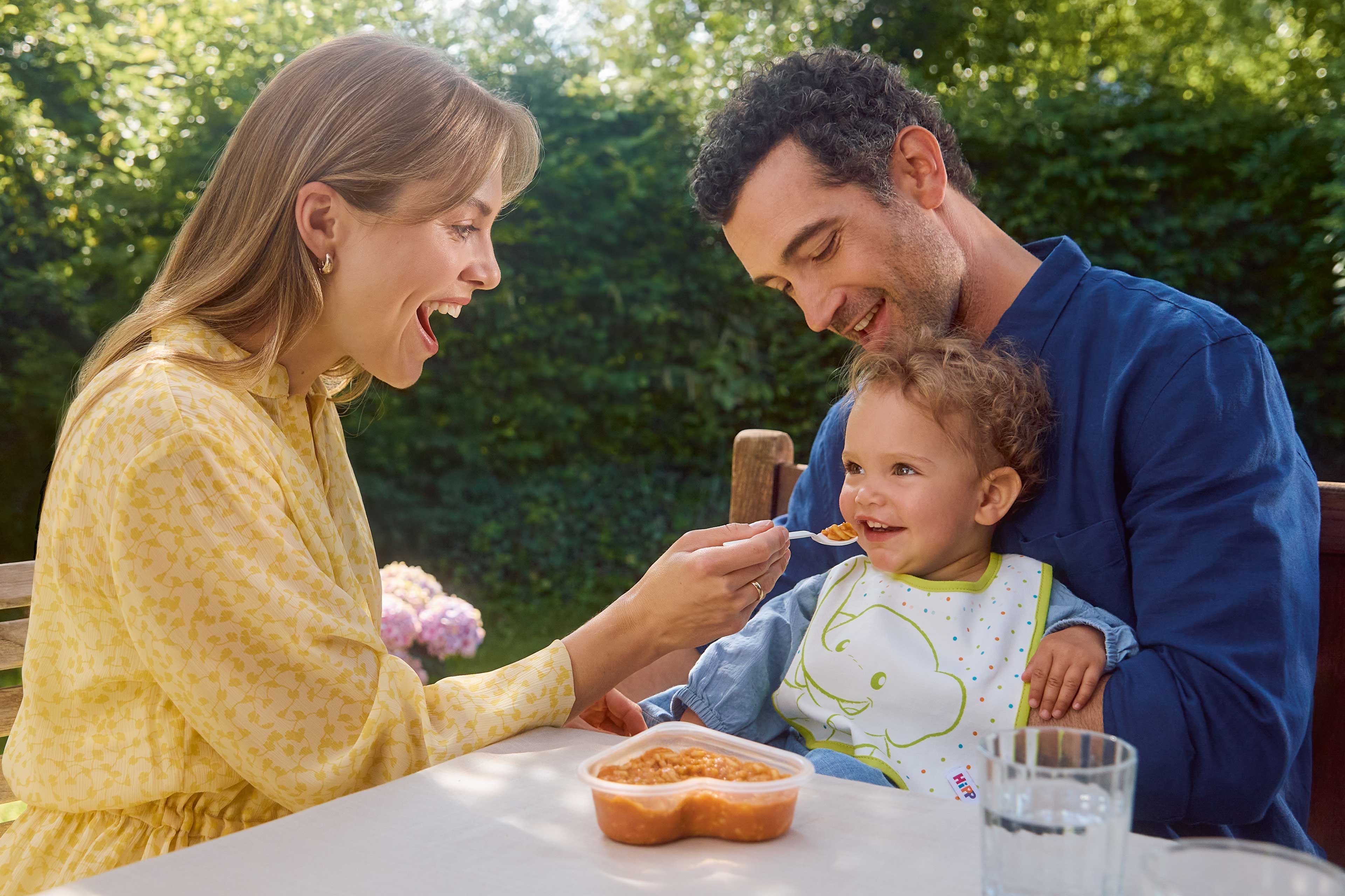 Eltern füttern ihr lachendes Kleinkind im Garten mit Beikostbrei – gemeinsame, entspannte Mahlzeit im Freien.