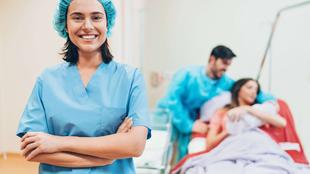 Medizinisches Personal in OP-Kleidung steht mit verschränkten Armen im Vordergrund, im Hintergrund sitzt eine Person auf einem Krankenhausbett und wird von zwei weiteren Personen betreut