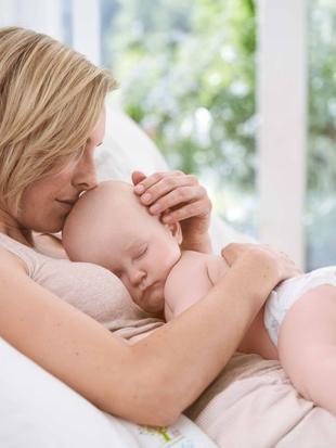 Eine Frau hält sanft ein schlafendes Baby, beide ruhen bequem auf einem Bett mit einem Fenster im Hintergrund.