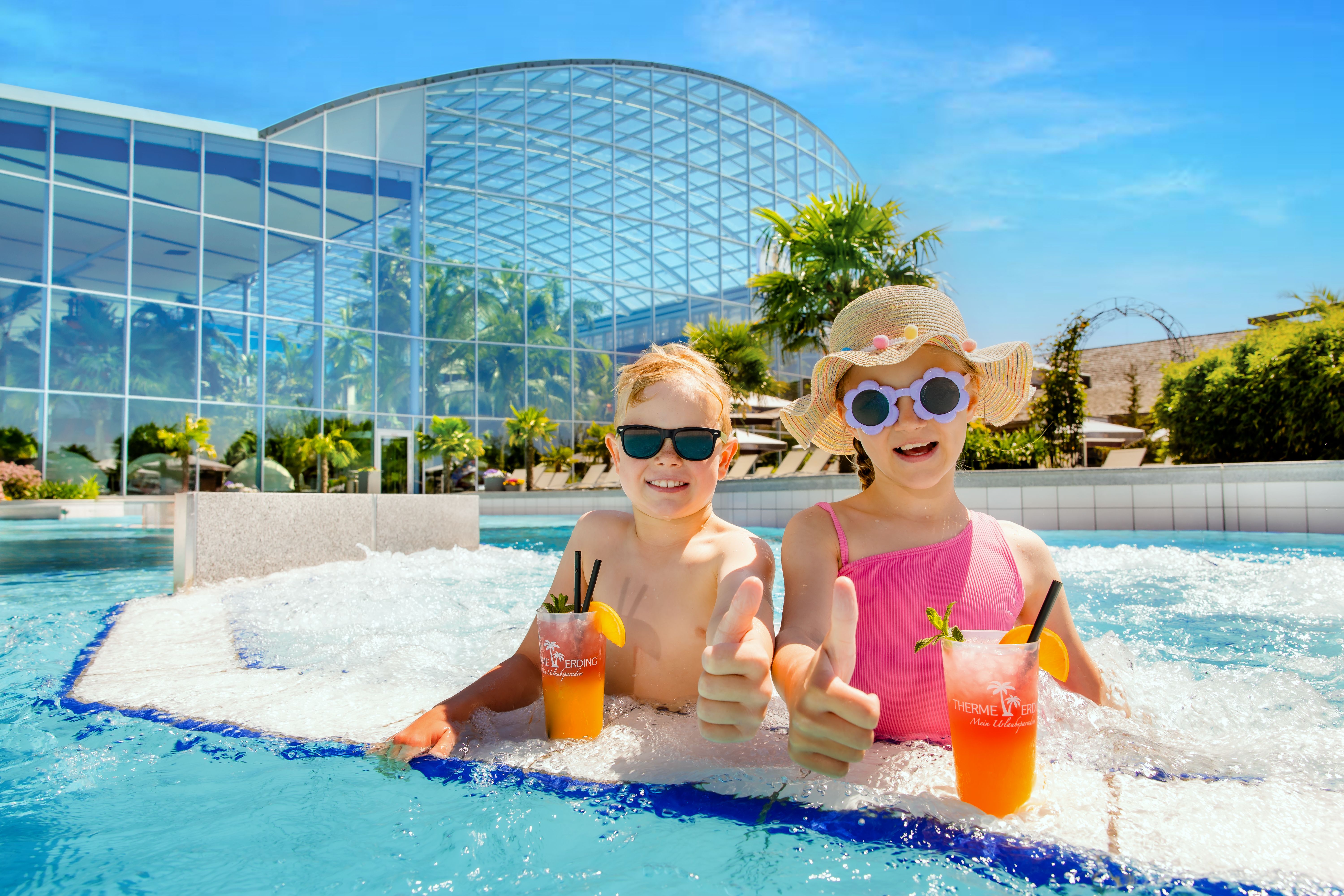 Zwei Kinder sitzen im Wasser mit einem Getränk und zeigen Daumen hoch