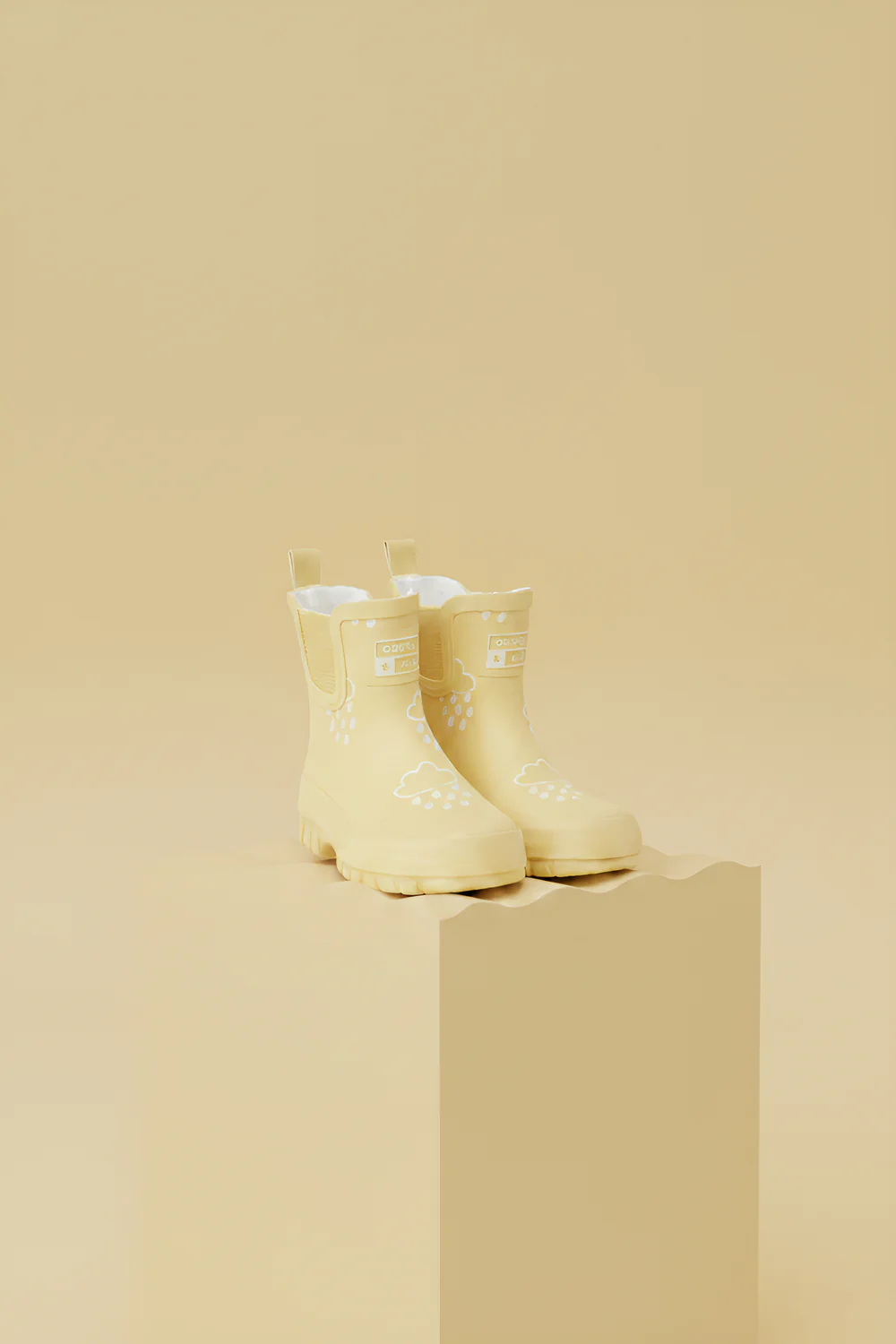 Pair of pale yellow Grass & Air children’s wellies with cloud and raindrop print on a matching beige background