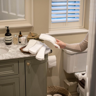 A person in a beige long-sleeve shirt holds a white reusable pad in a bathroom. Several other reusable pads are neatly placed on a wooden tray on the marble countertop, which also has bottles of soap, skincare products, and cotton swabs. The bathroom features a gray vanity, a toilet, and a small woven basket on the floor.