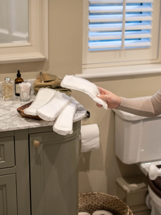A person in a beige long-sleeve shirt holds a white reusable pad in a bathroom. Several other reusable pads are neatly placed on a wooden tray on the marble countertop, which also has bottles of soap, skincare products, and cotton swabs. The bathroom features a gray vanity, a toilet, and a small woven basket on the floor.