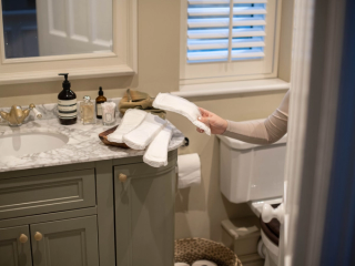 A person in a beige long-sleeve shirt holds a white reusable pad in a bathroom. Several other reusable pads are neatly placed on a wooden tray on the marble countertop, which also has bottles of soap, skincare products, and cotton swabs. The bathroom features a gray vanity, a toilet, and a small woven basket on the floor.