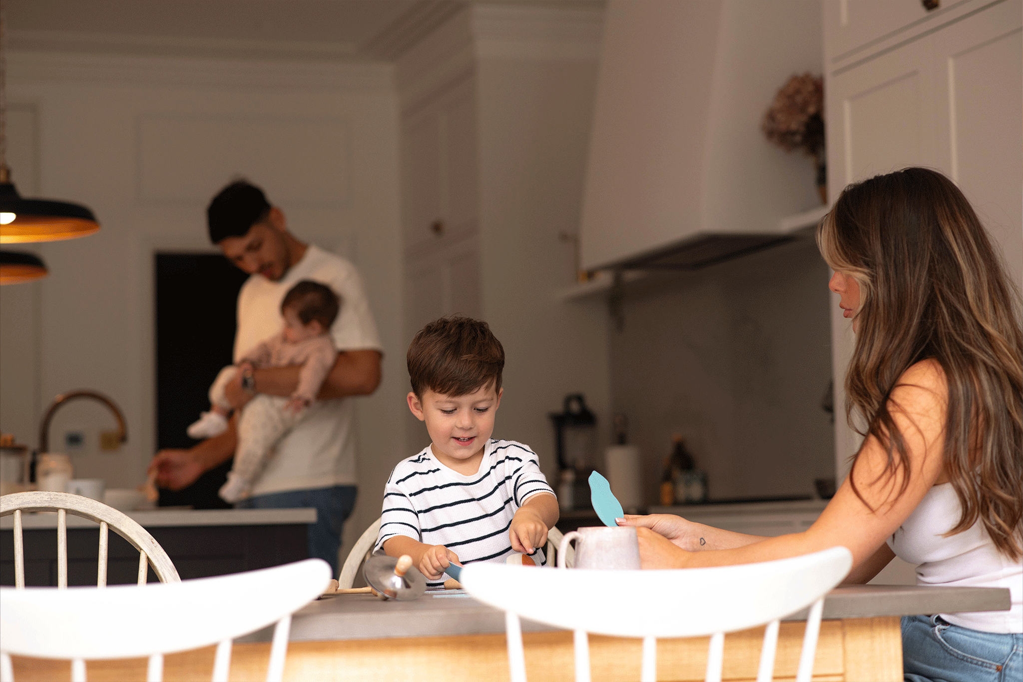 A wide shot of a modern kitchen scene featuring a family of four. In the foreground, a young boy in a striped shirt sits at the dining table engaged in an activity, while a mother with long brown hair sits opposite him, holding out a paper craft shape. In the softly focused background, a father stands holding a small baby near the kitchen island.