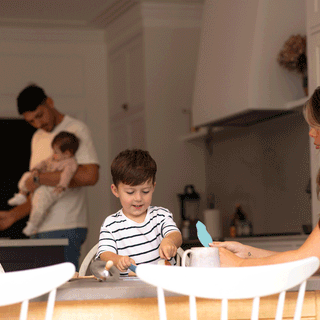 A wide shot of a modern kitchen scene featuring a family of four. In the foreground, a young boy in a striped shirt sits at the dining table engaged in an activity, while a mother with long brown hair sits opposite him, holding out a paper craft shape. In the softly focused background, a father stands holding a small baby near the kitchen island.