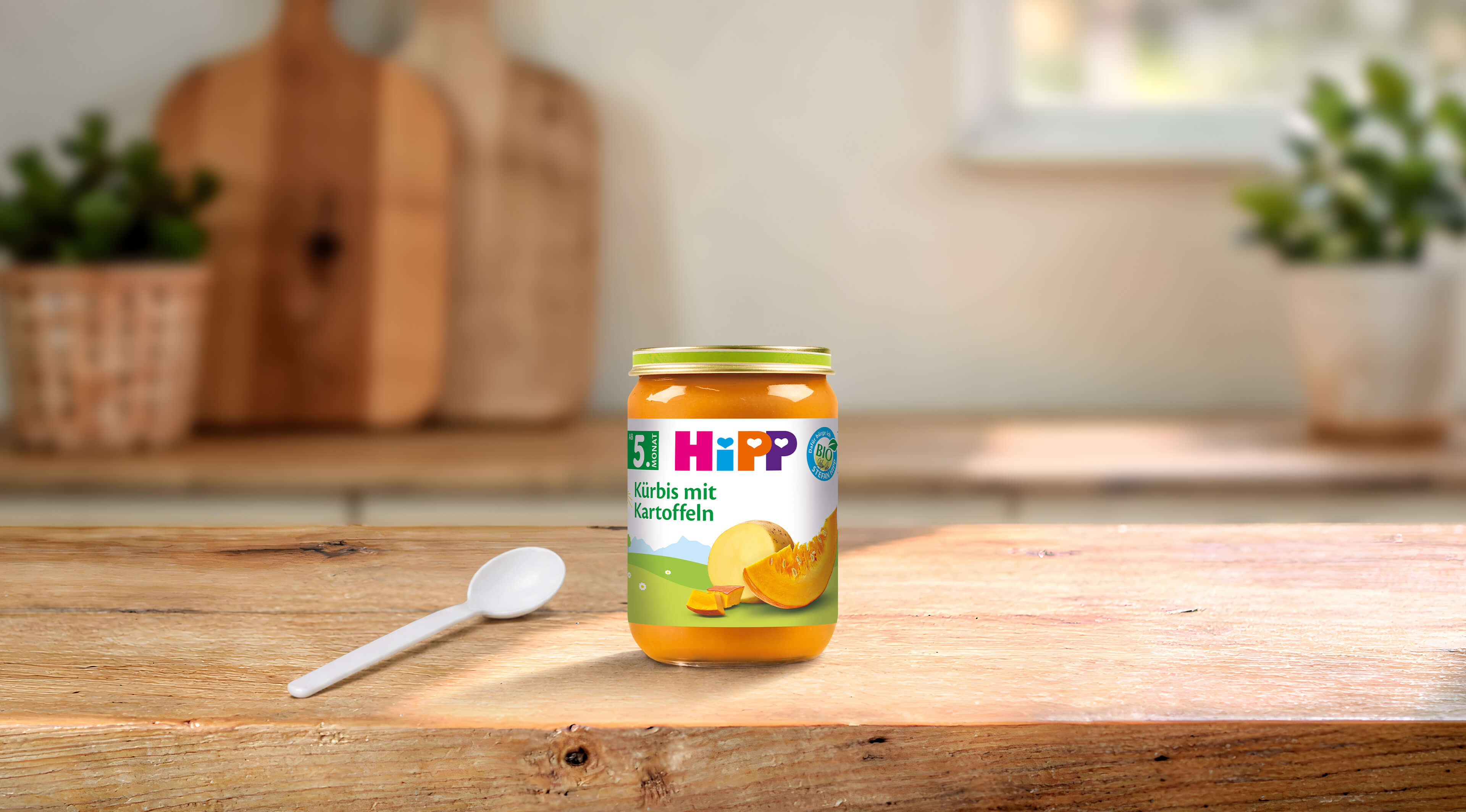 HiPP Bio-Gläschen „Kürbis mit Kartoffeln“ ab dem 5. Monat auf Holztisch, daneben ein weißer Babylöffel in heller Küche