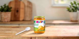 HiPP Bio-Gläschen „Kürbis mit Kartoffeln“ ab dem 5. Monat auf Holztisch, daneben ein weißer Babylöffel in heller Küche