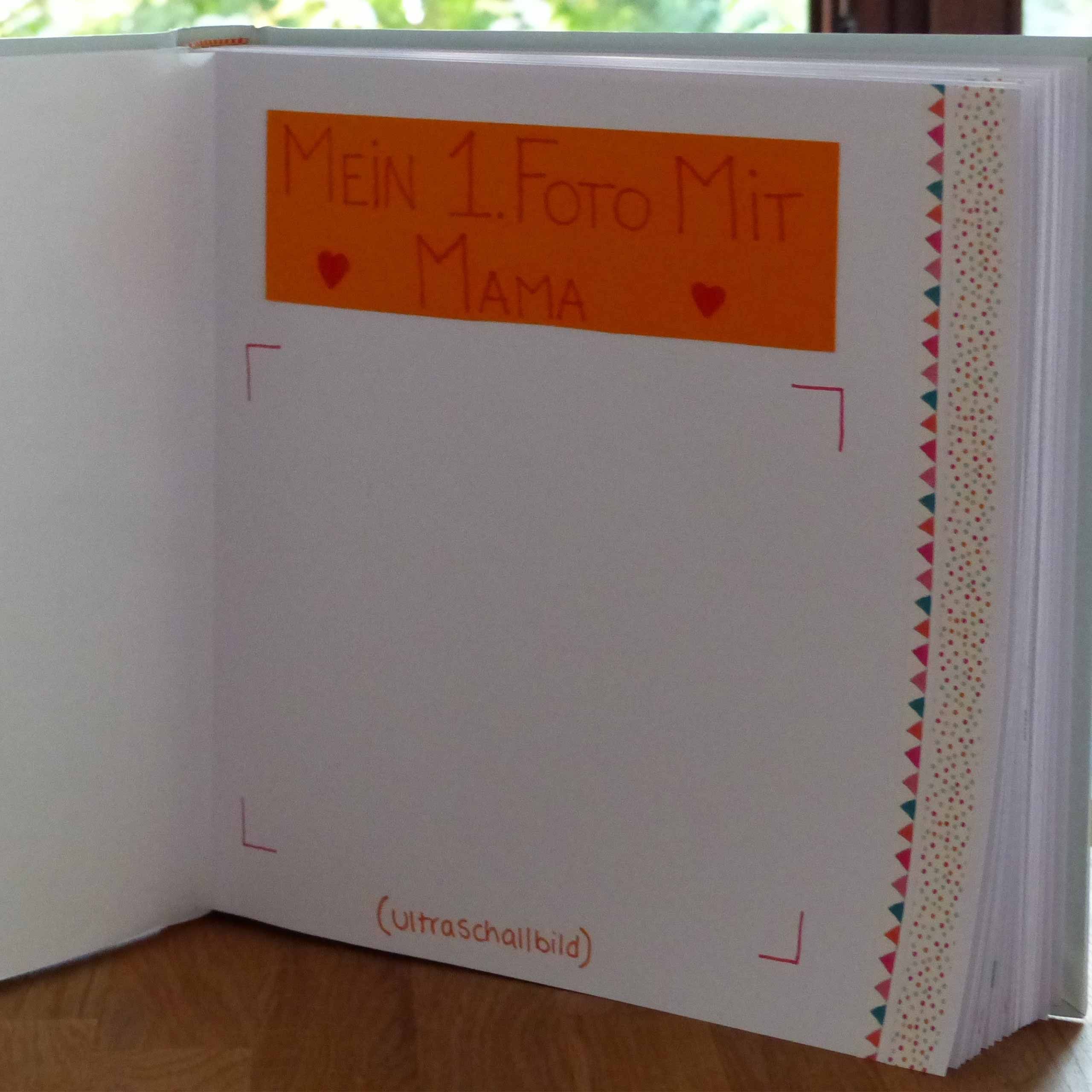Ein aufgeschlagenes Buch mit einer handschriftlichen Notiz auf einer orangefarbenen Seite, auf der "Mein 1. Foto mit Mama" steht, begleitet von zwei Herzsymbolen.