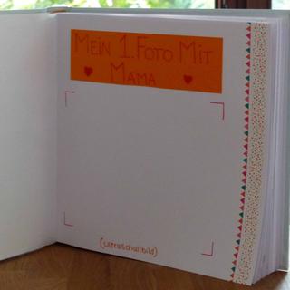 Ein aufgeschlagenes Buch mit einer handschriftlichen Notiz auf einer orangefarbenen Seite, auf der "Mein 1. Foto mit Mama" steht, begleitet von zwei Herzsymbolen.