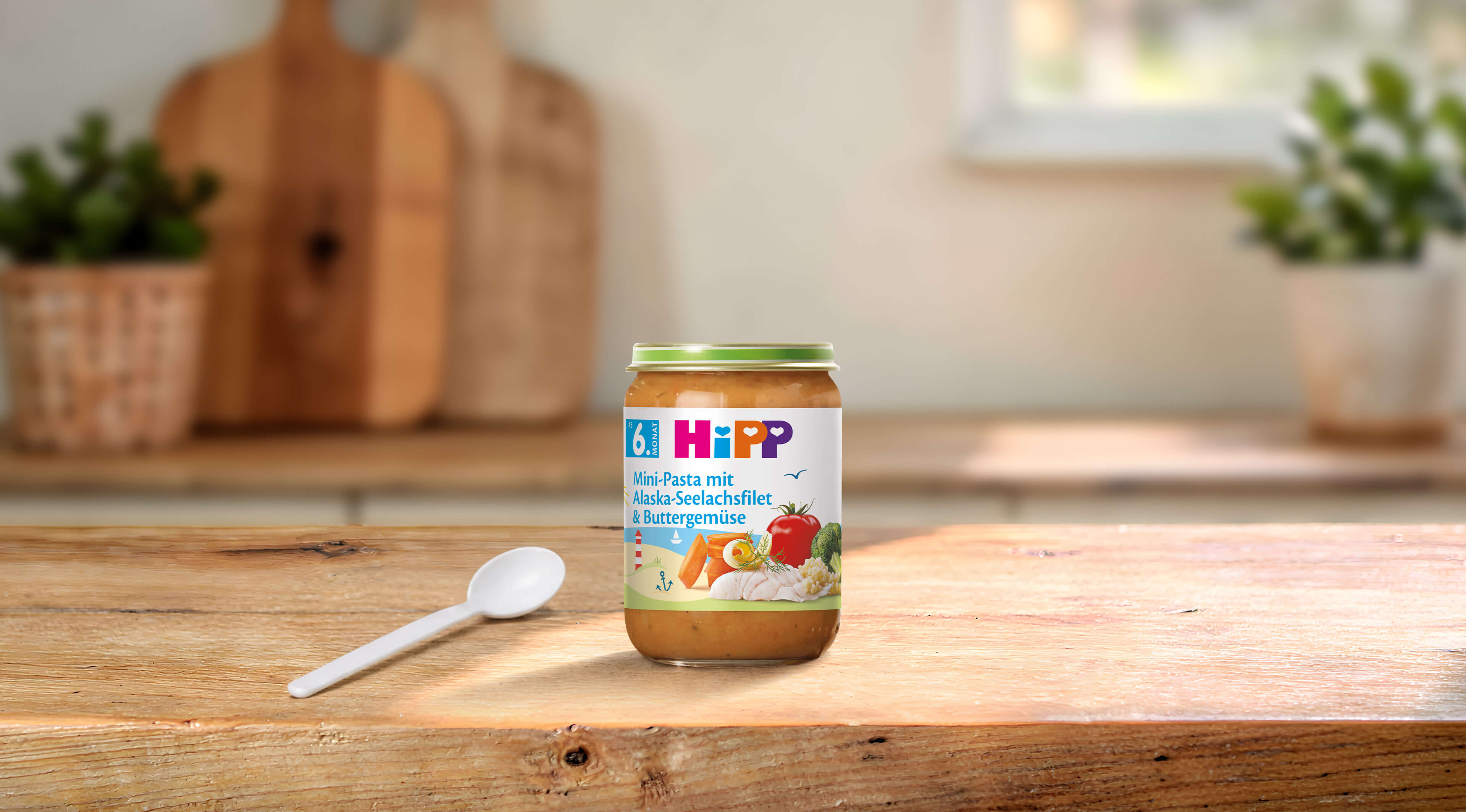 HiPP Mini-Pasta mit Alaska-Seelachsfilet & Buttergemüse im Glas auf Holztisch mit Löffel in heller Familienküche