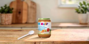 HiPP Mini-Pasta mit Alaska-Seelachsfilet & Buttergemüse im Glas auf Holztisch mit Löffel in heller Familienküche