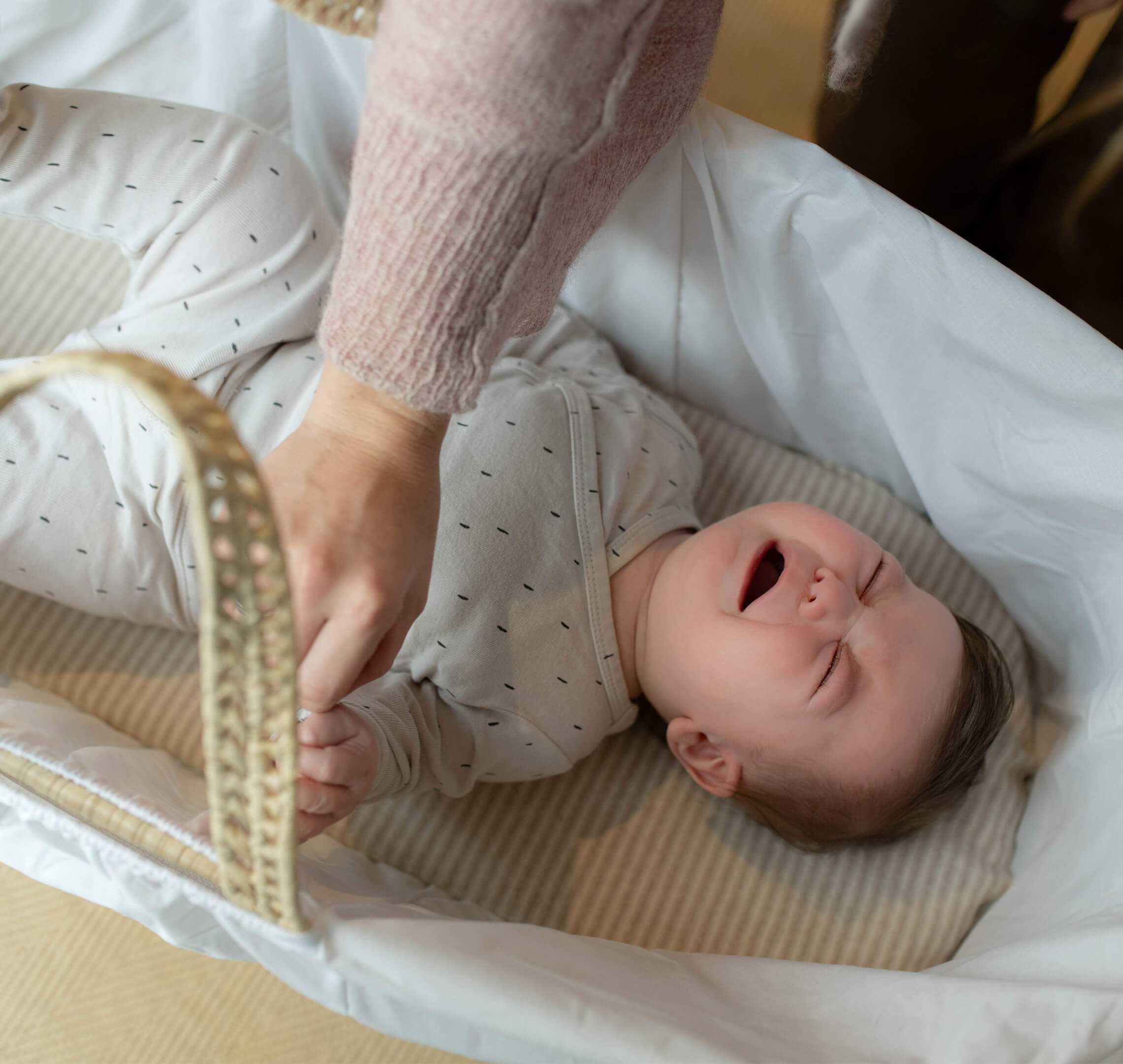 A baby lies in a white bassinet, crying with eyes shut tight and fists clenched, while an adult gently holds the baby's leg, possibly during a diaper change or soothing moment. The baby is dressed in a light onesie with a small dotted pattern.