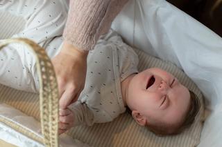 A baby lies in a white bassinet, crying with eyes shut tight and fists clenched, while an adult gently holds the baby's leg, possibly during a diaper change or soothing moment. The baby is dressed in a light onesie with a small dotted pattern