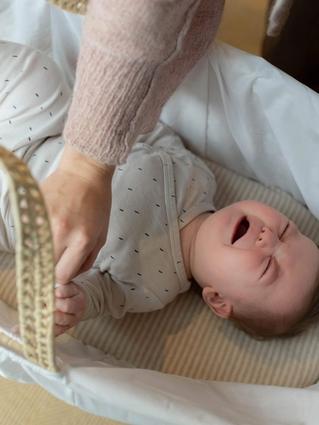 A baby lies in a white bassinet, crying with eyes shut tight and fists clenched, while an adult gently holds the baby's leg, possibly during a diaper change or soothing moment. The baby is dressed in a light onesie with a small dotted pattern.