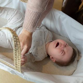 A baby lies in a white bassinet, crying with eyes shut tight and fists clenched, while an adult gently holds the baby's leg, possibly during a diaper change or soothing moment. The baby is dressed in a light onesie with a small dotted pattern.