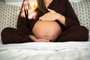 Close-up of a pregnant woman sitting cross-legged on a bed, gently cradling her bare baby bump with both hands. She is wearing dark brown silk pyjamas, with soft natural light shining across her.