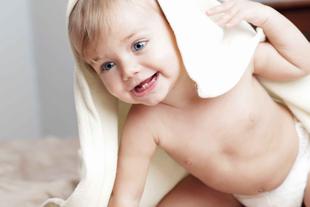 Ein fröhliches Baby mit blauen Augen, eingewickelt in ein weißes Handtuch, sitzt auf einer weichen Unterlage.