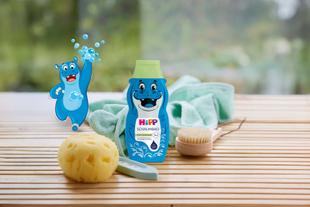 Flasche mit blauem Kinder-Schaumbad mit lachendem Nilpferd und grünem Deckel neben Schwamm, Bürste und gelbem Schwamm auf Holzuntergrund