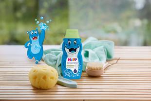 Flasche mit blauem Kinder-Schaumbad mit lachendem Nilpferd und grünem Deckel neben Schwamm, Bürste und gelbem Schwamm auf Holzuntergrund