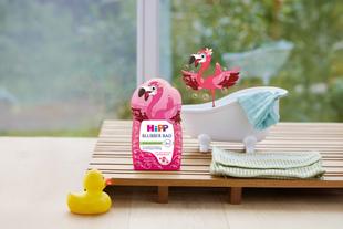 Flasche mit Flamingo-Motiv und der Aufschrift 'HiPP Blubber Bad' steht neben einer kleinen Badewanne mit Handtuch, einer gelben Quietscheente und einer gezeichneten Flamingofigur.