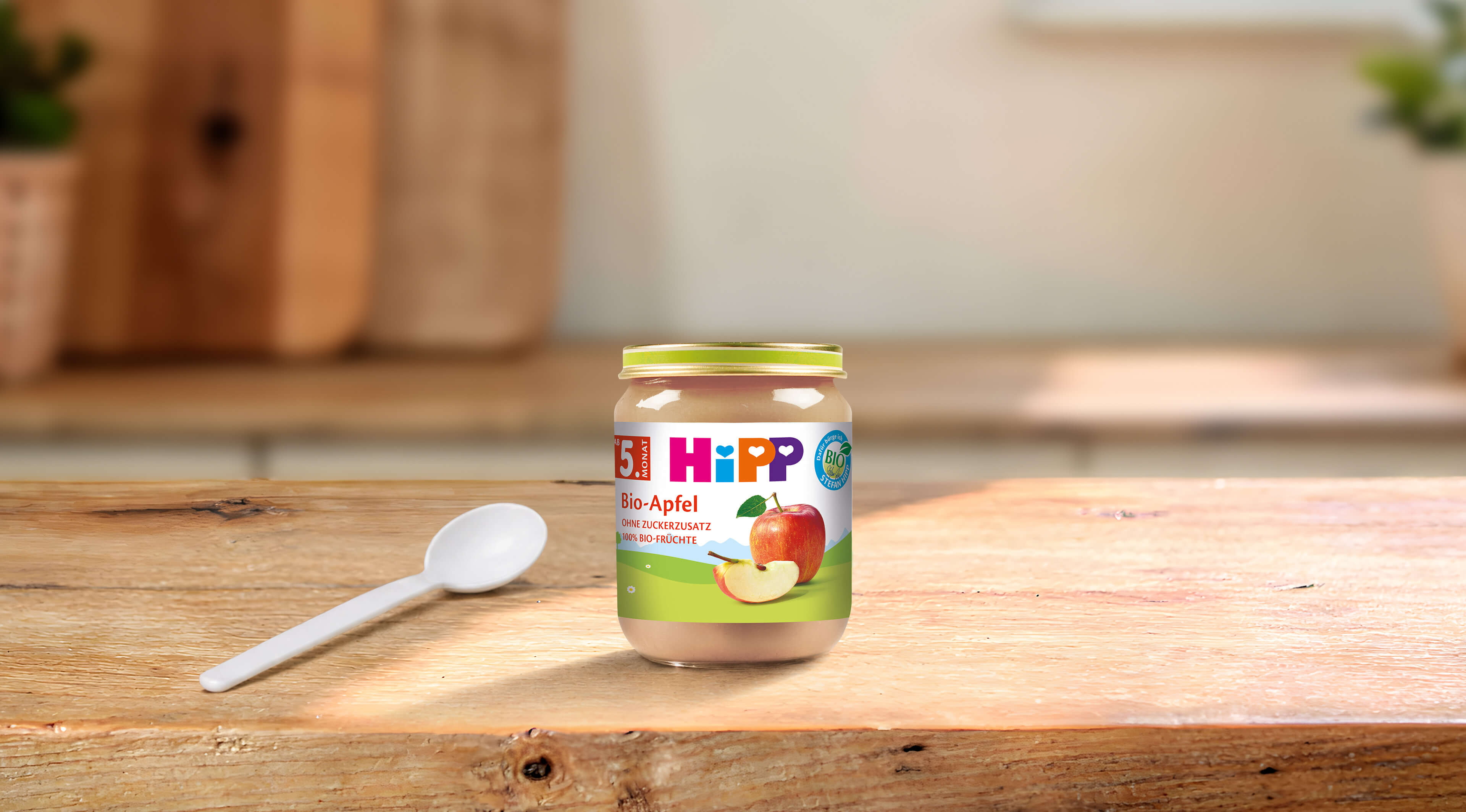 HiPP Bio-Apfel Babygläschen mit Apfelpüree und Löffel auf einem Holztisch in heller Küchenumgebung