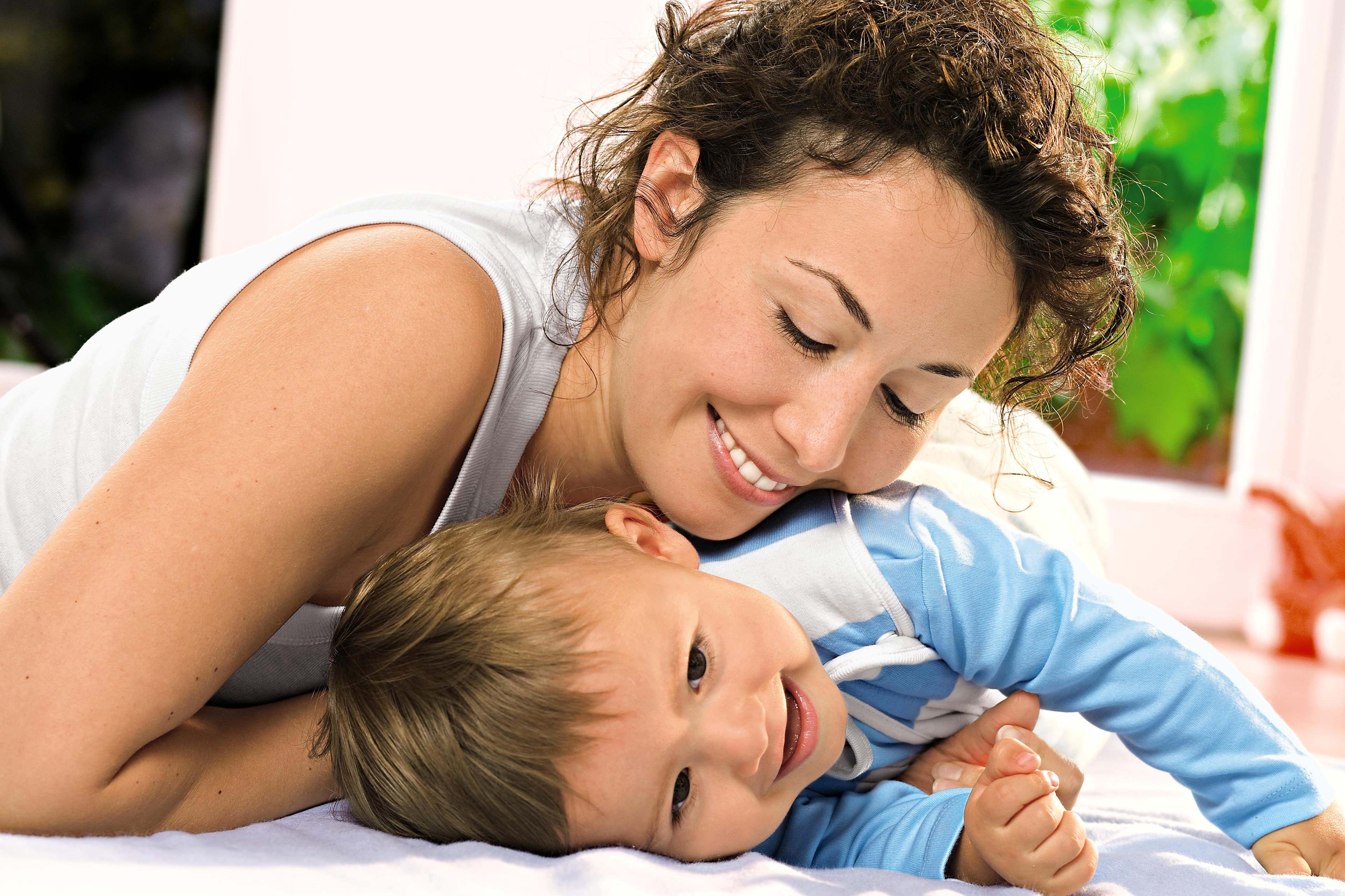 Lächelnde Mutter kuschelt sich zu ihrem Baby auf dem Bett, beide wirken entspannt und geborgen.
