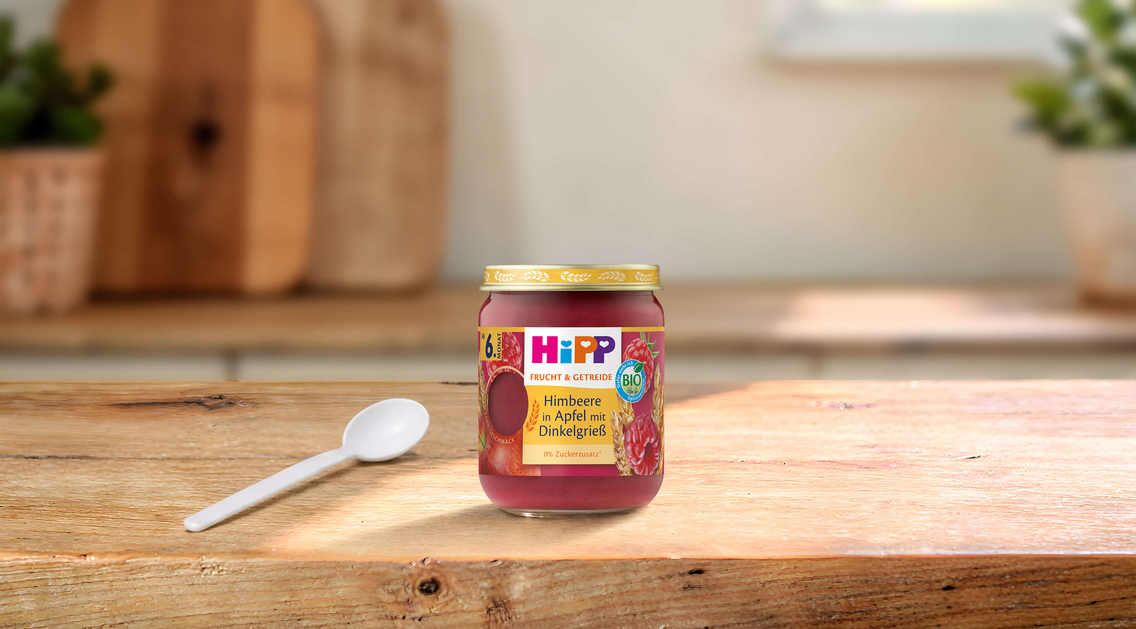 HiPP Bio-Gläschen „Himbeere in Apfel mit Dinkelgrieß“ ab 6. Monat auf Holztisch, daneben ein weißer Babylöffel