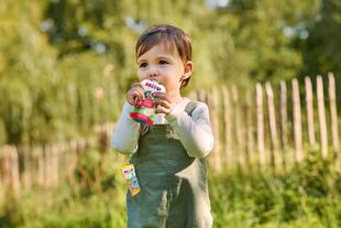 Kleinkind im Garten trinkt einen HiPP Bio-Quetschie und hält einen weiteren Kindersnack in der Latzhosentasche