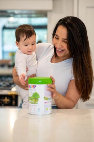 Mum holding baby touching a follow on milk tin while leaning on a kitchen worktop