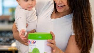 Mum holding baby touching a follow on milk tin while leaning on a kitchen worktop