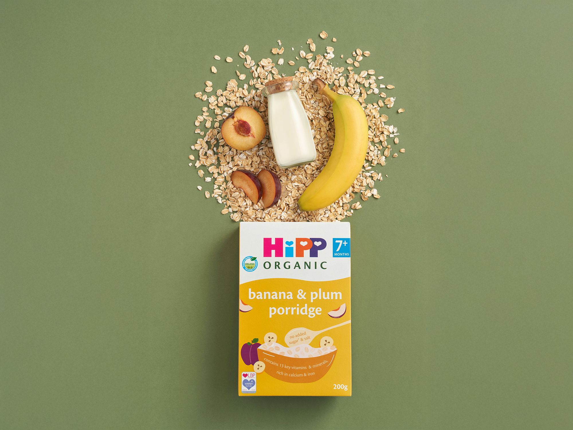 HiPP Organic banana & plum porridge box with oats, banana, plum slices and milk arranged on a green background