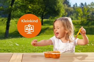 Kleinkind sitzt draußen an einem Holztisch und hält einen Löffel über eine orangefarbene Brotdose