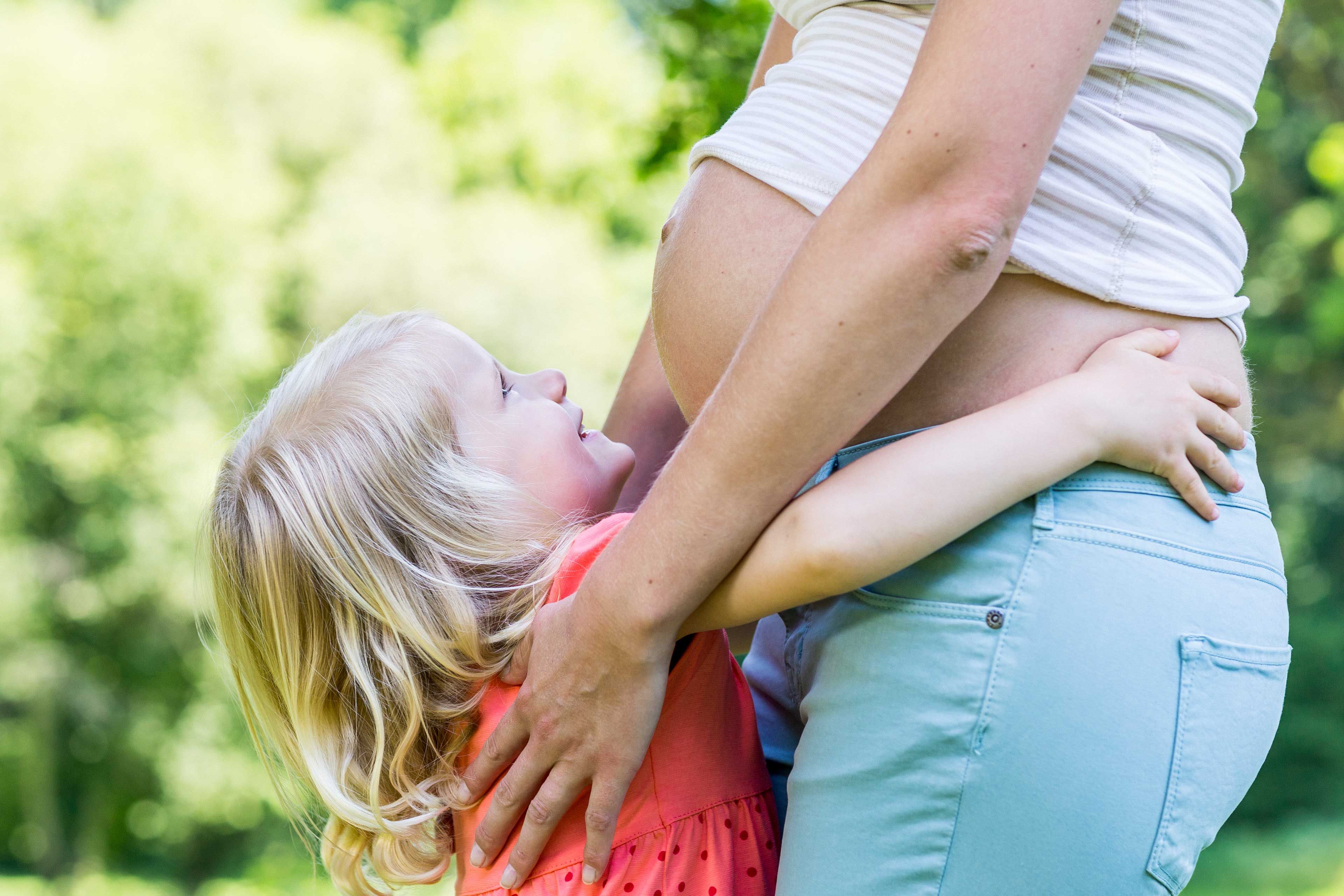 Schwangere Frau im Profil mit sichtbarem Babybauch umarmt ein kleines Kind im roten Kleid im Freien