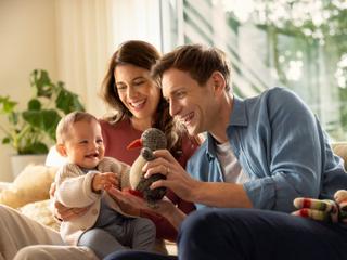 Lachende Eltern spielen auf dem Sofa mit ihrem Baby und einem Stofftier in einer warmen, hellen Wohnzimmeratmosphäre.