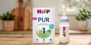 HiPP 1 PUR Bio-Anfangsmilch Packung neben gefülltem Babyfläschchen auf Holztisch in heller Küche