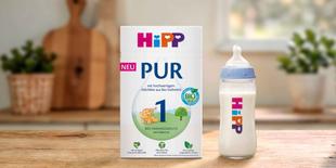 HiPP 1 PUR Bio-Anfangsmilch Packung neben gefülltem Babyfläschchen auf Holztisch in heller Küche