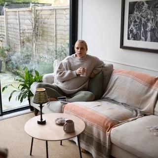 person sits cross-legged on a cream-colored sectional sofa with light pink and beige throws, looking relaxed in a cozy, modern living room. Large glass sliding doors open to a small patio garden, and a framed black-and-white artwork hangs on the wall above the couch.