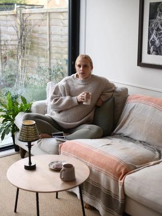 person sits cross-legged on a cream-colored sectional sofa with light pink and beige throws, looking relaxed in a cozy, modern living room. Large glass sliding doors open to a small patio garden, and a framed black-and-white artwork hangs on the wall above the couch.