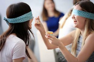 Zwei Frauen, eine mit einem blauen Stirnband, füttern die andere mit einem Löffel, während beide lachen und lächeln.